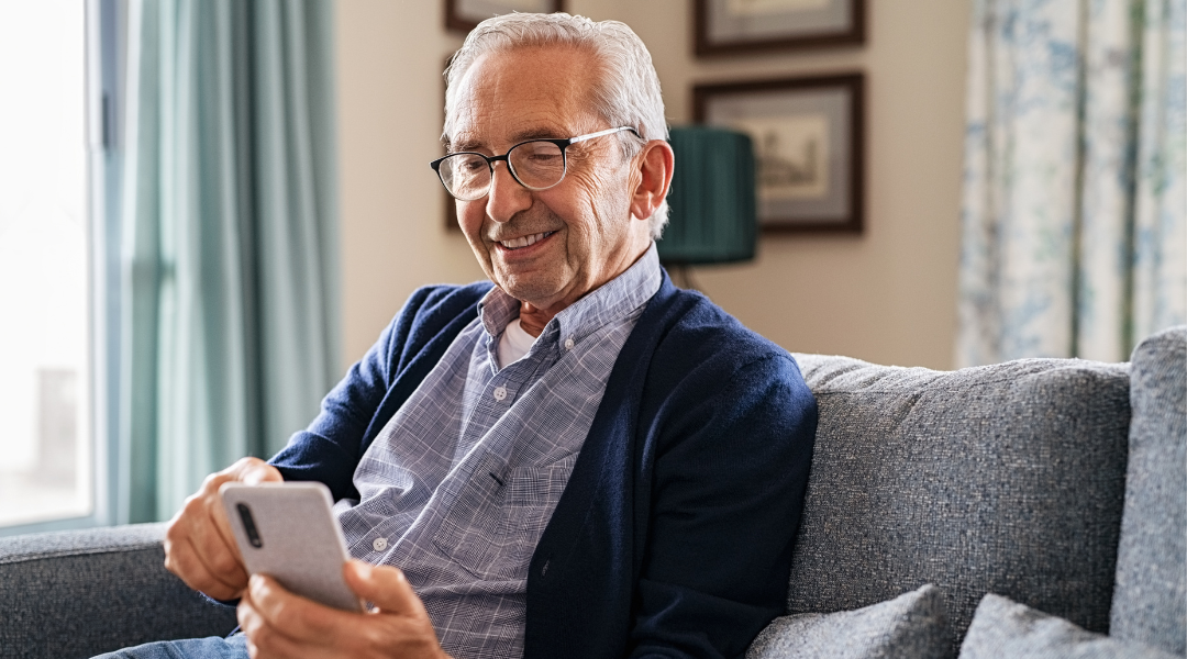 Smartphonekurs für Senioren Ein grauhaariger Mann in Jeans und mit Brille sitzt auf einem Sofa. Er schaut in sein Smartphone und lächelt.